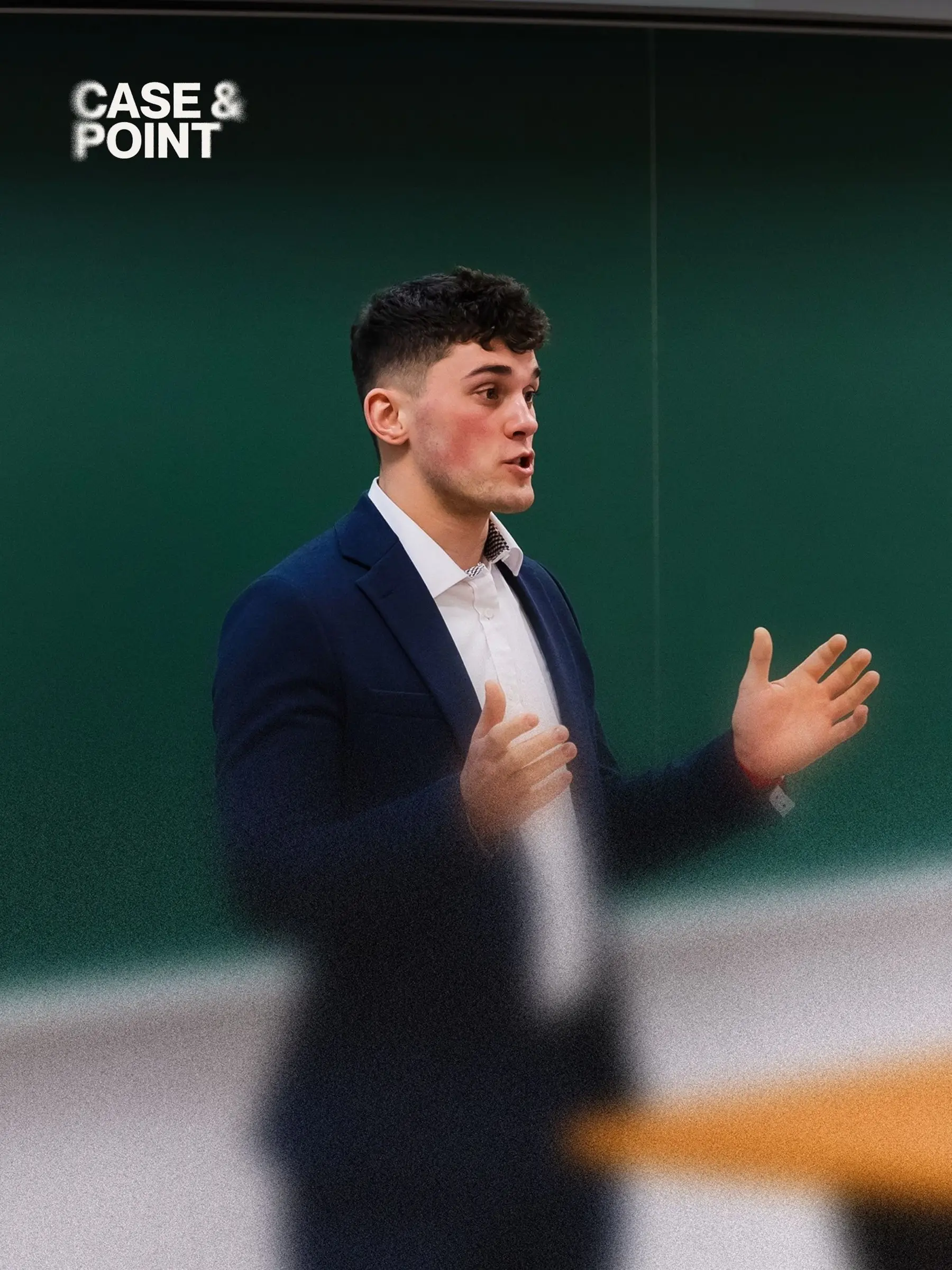 A JMCC delegate in a navy blazer presenting to a room, mid-gesture, in front of a green chalkboard